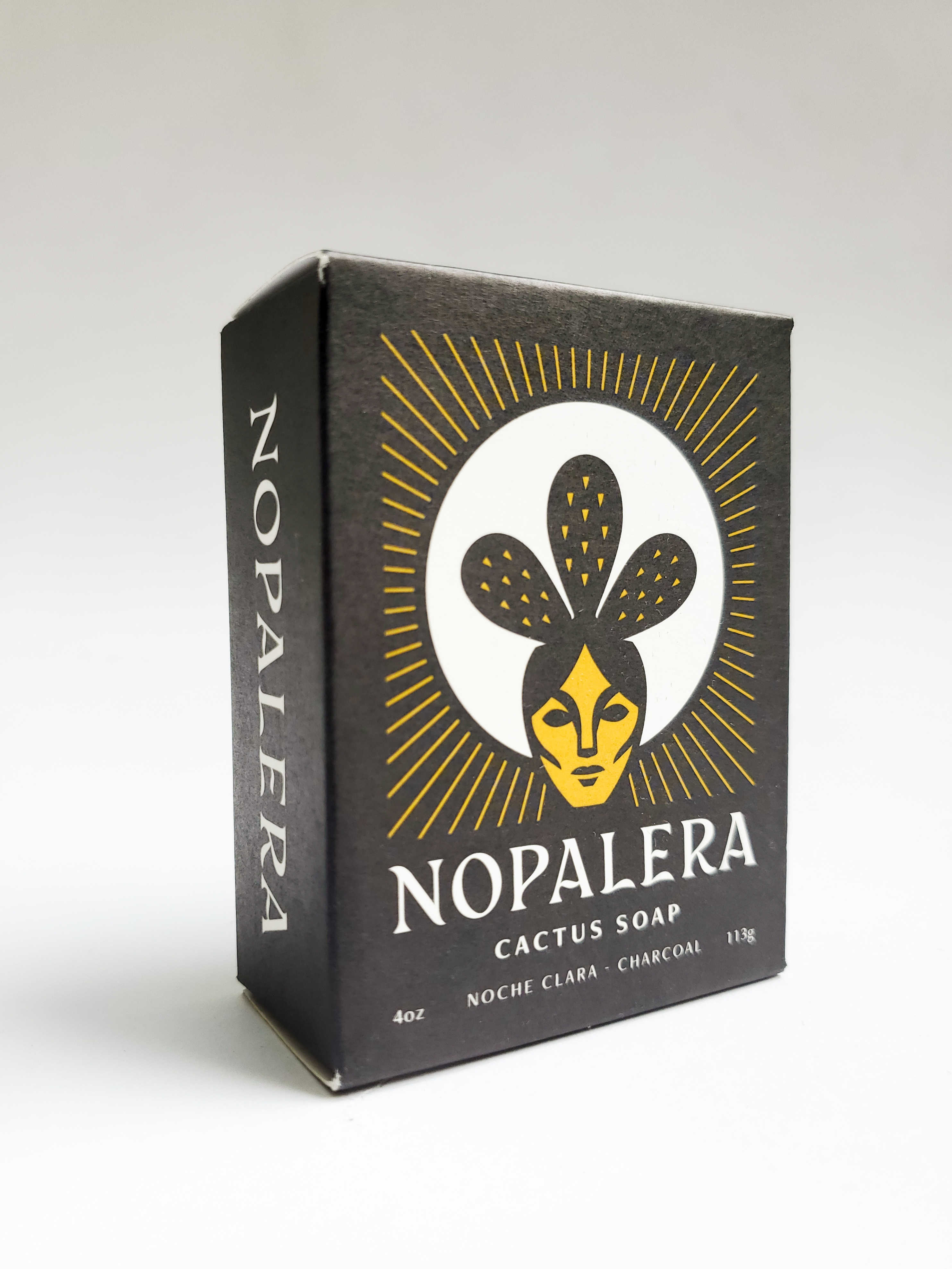 A black box with an image of a woman with a cactus plant on top of her head, standing in front of the sun. Text on the box says "Nopalera Cactus Soap. 4 oz. Noche Clara. Charcoal. 11%."