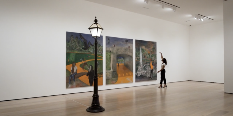 Lightpole in front of a triptych. One woman stands to the right of the paintings with one arm raised