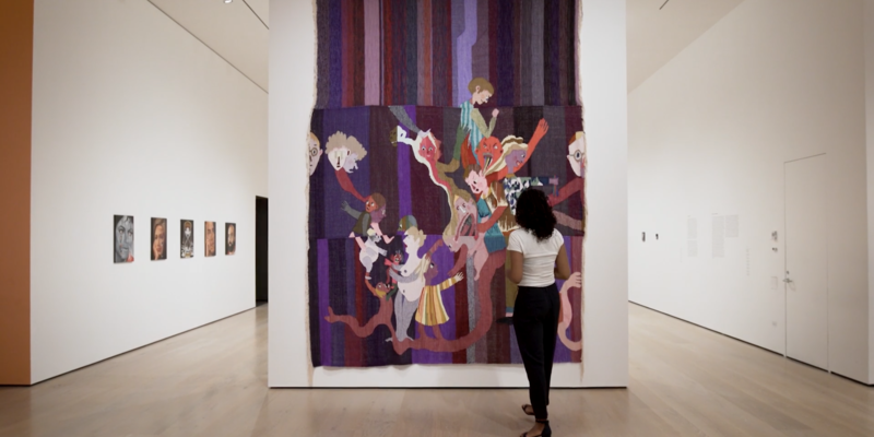 A woman stands center right in front of a multi-colored tapestry with a purple background and many human-like figures in the foreground