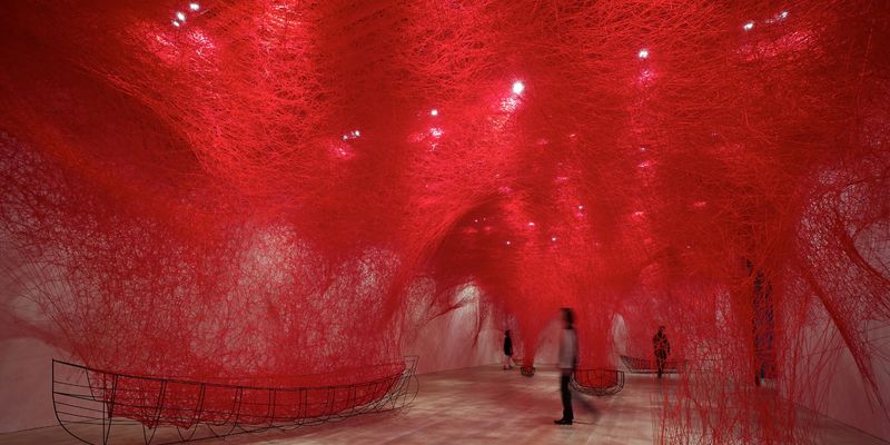 A person walking under an installation of fine red thread suspended from the ceiling and connect to small boat frames