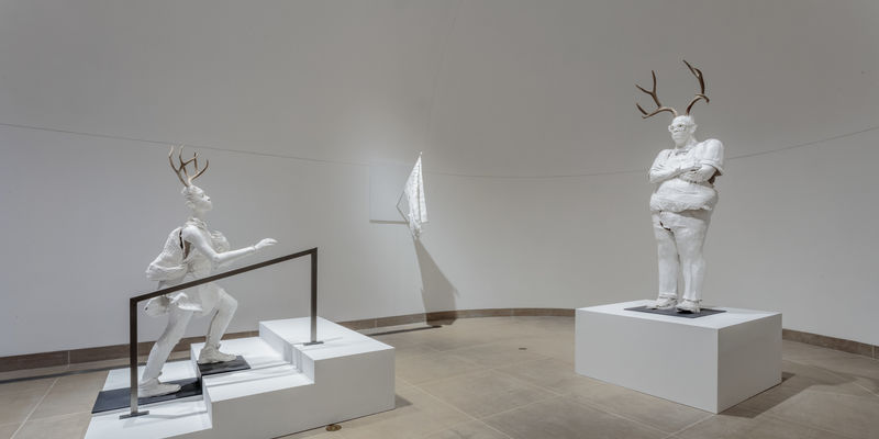 Life-sized white plaster figures of a girl climbing the stairs and a principal with arms crossed, both with antlers on their heads 