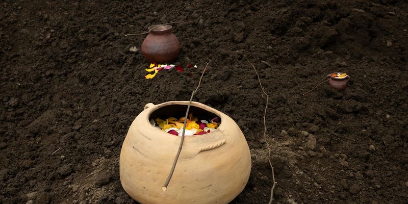 Three earthen pots, filled with flower petals with a twig laying across the top, on a bed of soil