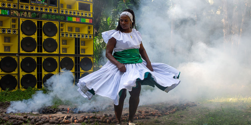 A woman in a white dress dances in front of a wall of yellow speakers, surrounded by smoke