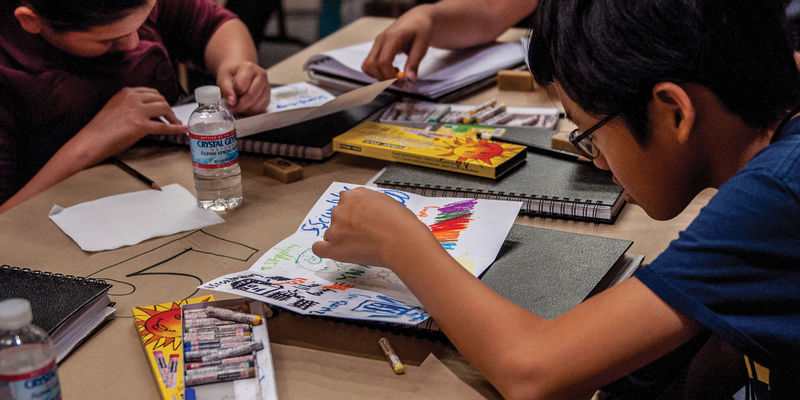 Two children sit across a table and draw on paper