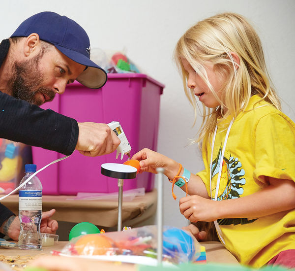 A girl in a yellow shirt is holding up an art project and is assisted by a man in a baseball cap who leans over with a glue gun pointed at the girl's project.