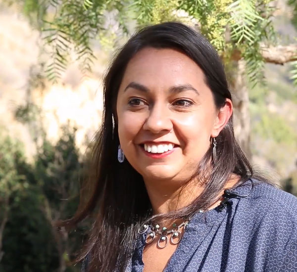 Photo of artist Mercedes Dorame, a woman with long brown hair smiling at someone standing to left of cameral.   She is outside, with a hillside of foliage behind her. She wears a blue shirt and a silver necklace.