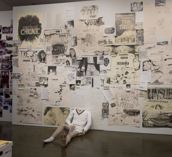 A gallery wall covered in drawings, with a headless mannequin sitting against it