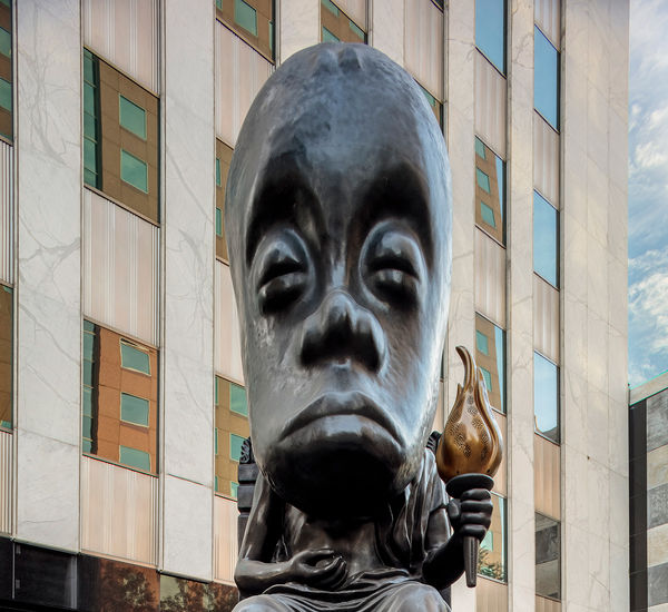 Large-scale public sculpture of a seated oracle figure holding a torch