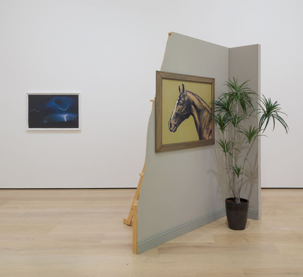 installation image with framed work on wall and a sculpture comprised of a false wall with painting and potted plant