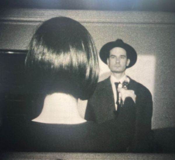 Black-and-white image of the back of a person's head with short black hair, pointing a metal object at another figure wearing a black suit and hat