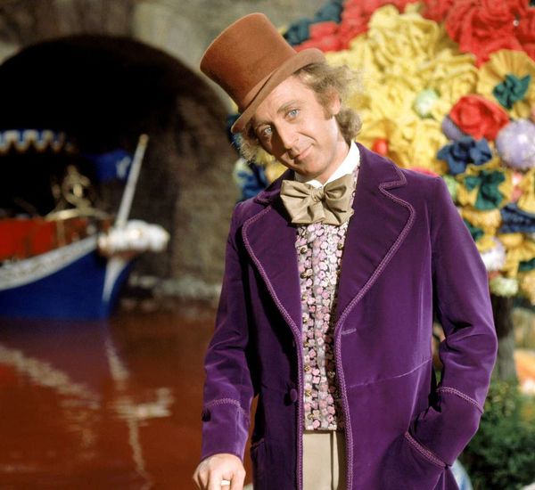 Willy Wonka wearing a brown top hat and purple coat stands in front of his chocolate river