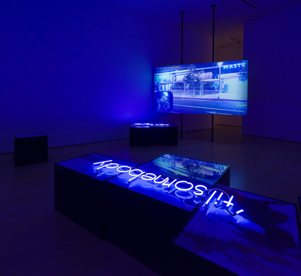 Installation with a video screen in the background showing a street scene, and a neon sign reading "til' somebody" laying on a plinth in the foreground