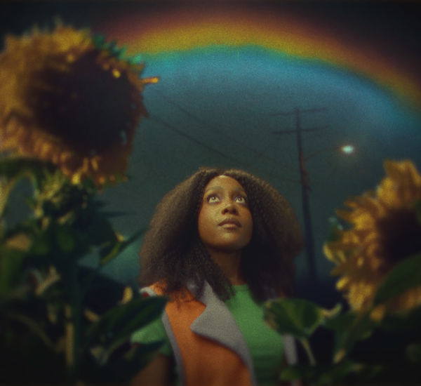 A Black woman, framed by sunflowers, looks up at a rainbow in the night sky