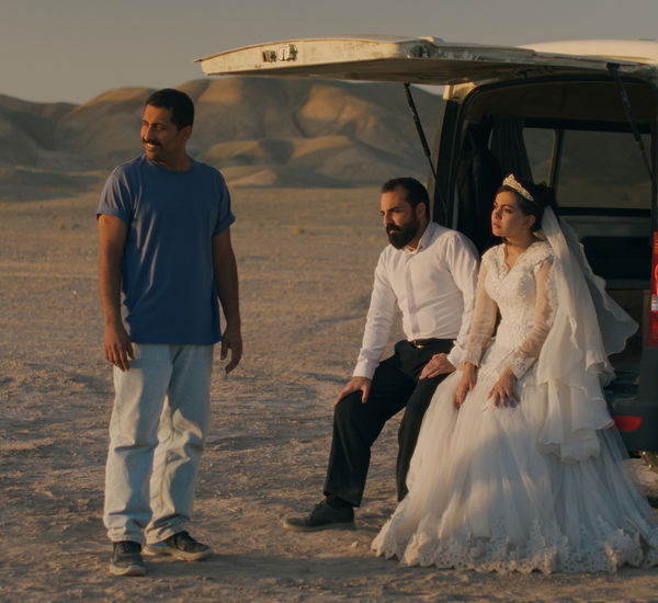 A couple dressed for their wedding sits in the open hatch of a van in the dessert