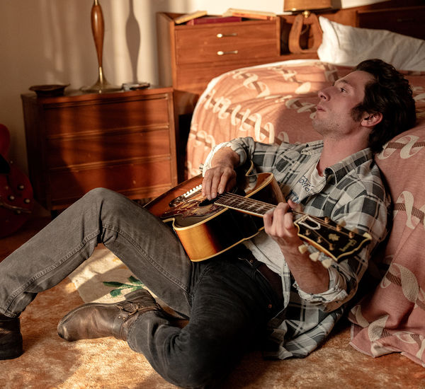 Jeffrey Allen White as Bruce Springsteen sits against a bed playing an acoustic guitar