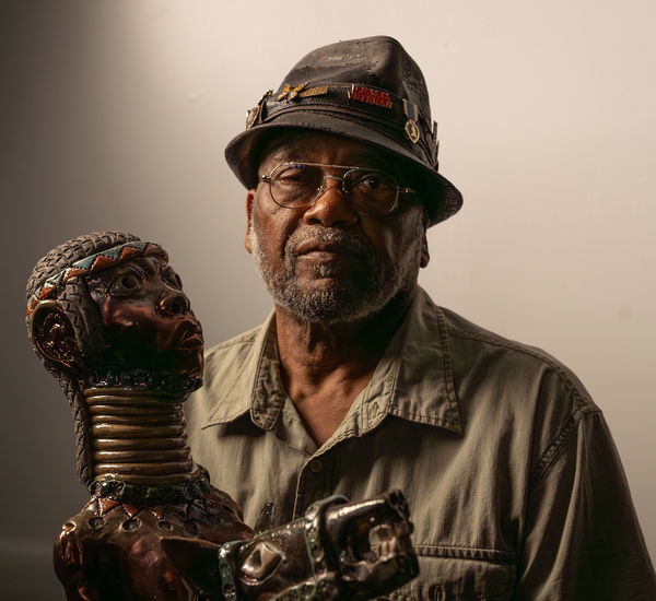 The artists Akinsanya Kambon sits next to his sculpture of a long-necked figure
