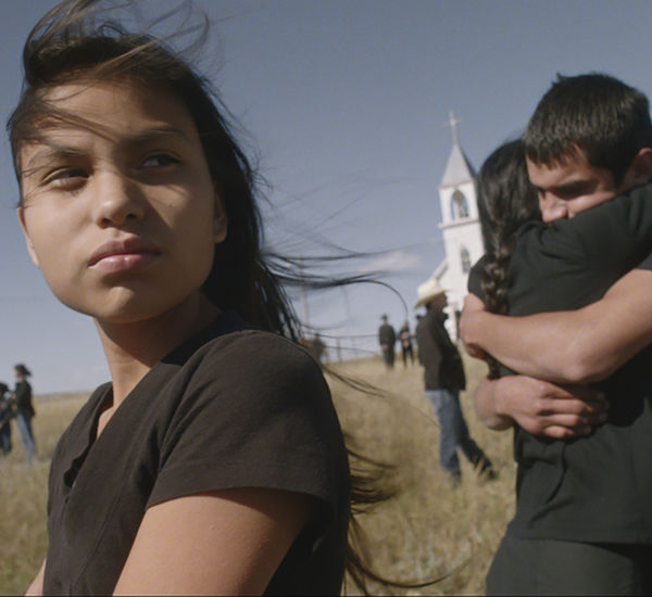 A girl looks off-camera, while behind her a man and woman hug in front of a church
