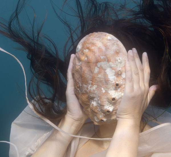 A woman, underwater, holds a large shell in front of her face