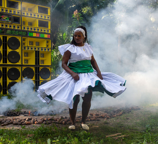 A woman in a white dress dances in front of a wall of yellow speakers, surrounded by smoke