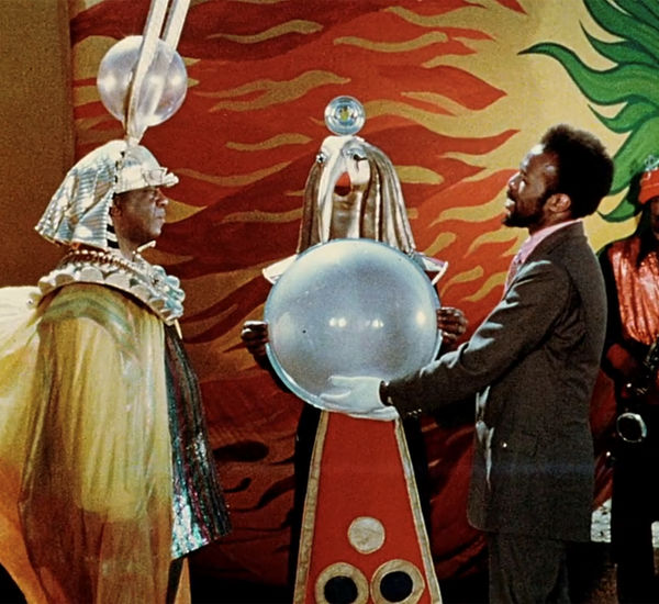Sun Ra, in an elaborate headdress, stands with a man holding a large orb in his white-gloved hands