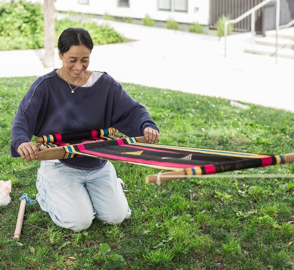 A woman kneels on the ground, holding a weaving suspended in front of her