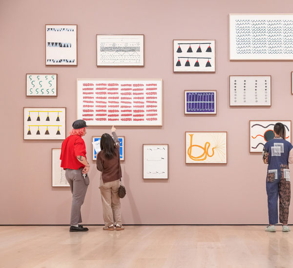 Three visitors, viewed from behind, stand in front of a pink gallery wall covered in a cluster of framed abstract artworks