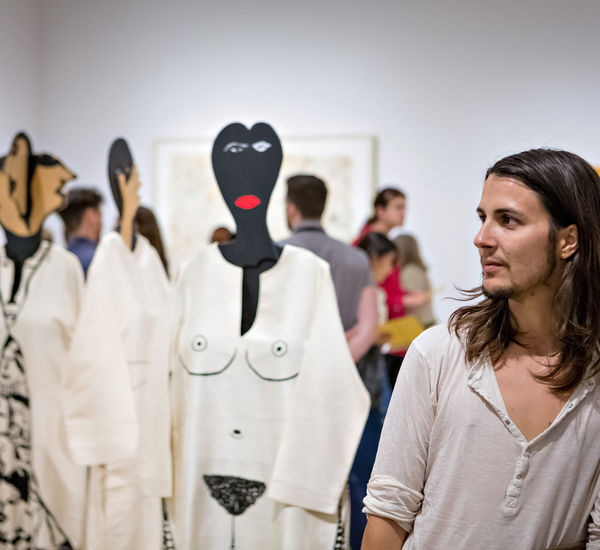 A person with long, straight brown hair and a short beard glances to the right. Just behind him are visible four mannequins wearing caftans with designs painted onto them