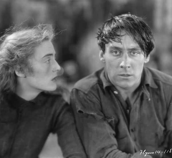 On the right, a man with dark hair swept across his forehead looks at the camera, while a woman on the left gazes at him