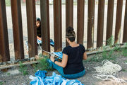 Two women sit on either side of the US-Mexico border fence, connected by a woven rope between them