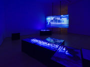 Installation with a video screen in the background showing a street scene, and a neon sign reading "til' somebody" laying on a plinth in the foreground
