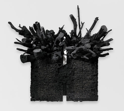 a three-dimensional, black rectangular shape hangs on a white wall, with black sticks sticking out from the top. The appearance is of charcoal stacked and sticks that look blackened with charcoal.