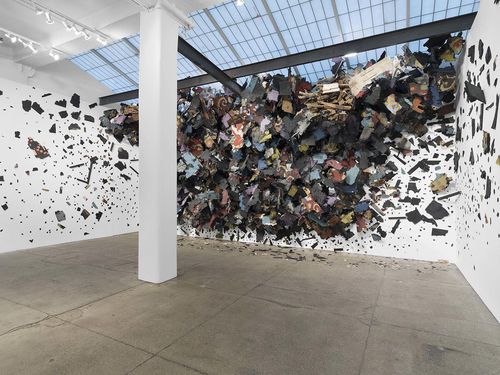 A view of a room with white walls has debris covering the far wall and appearing to float out in front of the wall. The room has clearstory windows on the ceiling. 