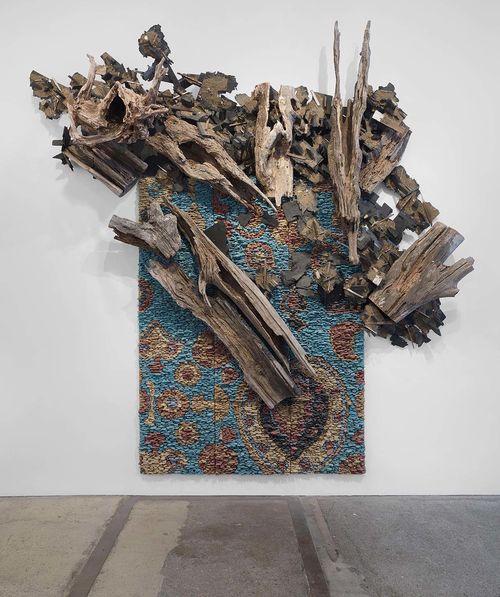 A rectangular work of art hangs on a white wall. The work has a textured surface that appears to be stacked with colored pieces of wood or stone that are blue, tan and brown. Pieces of deteriorated wood are attached to the surface, mostly near the top of the work, and stick out beyond the rectangle. 