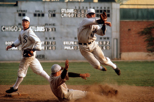 Still from the film Eight Men Out (1988) showing a baseball player slide into a base while another player, on the White Sox team, jumps and throws a ball to complete a double-play while a third player looks on