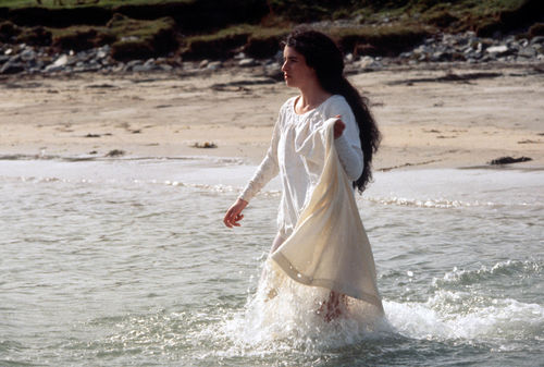 Still from the film The Secret of Roan Inish (1994) showing a woman in a white dress wading into the water on a beach shore