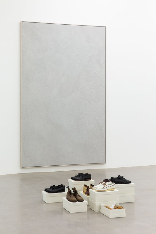 Shoes on top of boxes are on the floor before a gray square on the wall