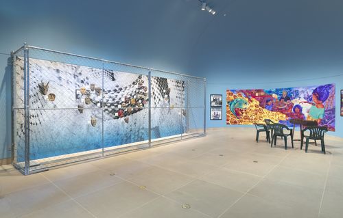 A chainlink fence around a display of masks, with a colorful mural hung on the wall next to them and green chairs on the gallery floor