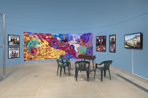 A colorful mural against a pale blue wall with displays on either side, and green chairs on the gallery floor