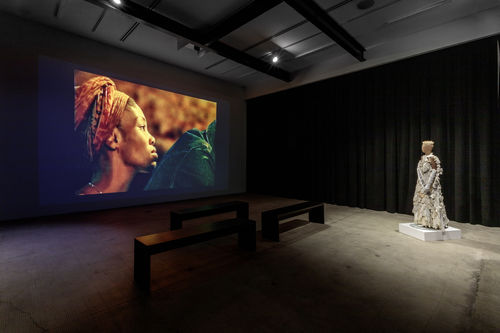 A dark gallery with a projection on one wall and a costume displayed near another