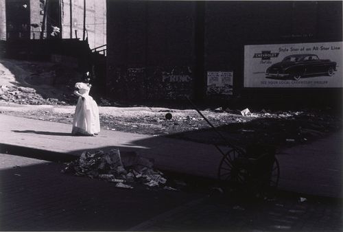 A young woman in a white dress walks through rubble