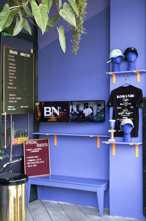 Photo looking inside a room with purple wall and slanted roof. Against the purple wall are two video monitors with BN letters in white displayed on the left and a man n white tshirt on the right. In the room hats and a tshirt are displayed on shelving.