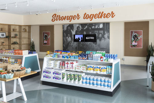 Photograph showing inside of a grocery store, with low shelving. On the back wall the words "Stronger Together" are written in orange cursive. Below this a black and white photographic mural, with two video monitors mounted over top.