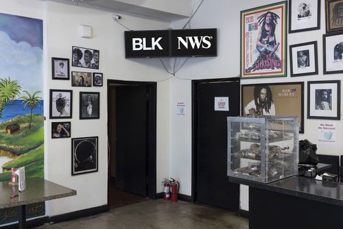 Photograph of a corner of a large room with many framed images on the walls. The walls are white. The corner of the room is in the center of the photograph and two dark doors are on either side of the corner. Above the doors are two video monitors with white letters BLK and NES on each monitor.