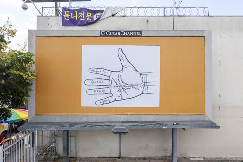 Photo of a billboard against a white building, with a square image of a palmistry diagram in black and white  pasted on top of a gold backgrounf.