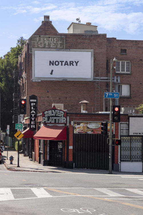 Image of a brick building on a street with commercial businesses on street level. On the side of the building is a billboard with black letters "NOTARY" written on a a white background and an arrow pointing toward he street.