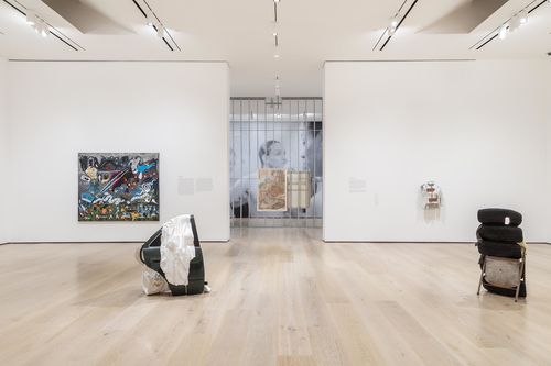 Three sculptures made of mixed objects are displayed prominently in a gallery. Two are on the floor in the middle of the gallery and a third hangs on a wall to the right. A painting hangs on a left. A passageway splits these two walls into another gallery.