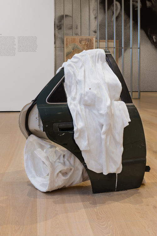 An image of a sculpture with two car doors propped up vertically. Extending from the floor and spilling out of the car door window is a white sheet made of plaster. 