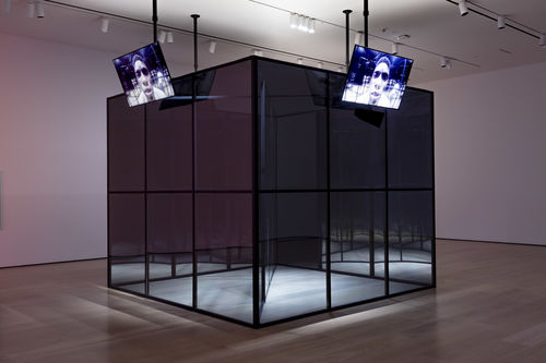 A large, mirror-like cube in the middle of a gallery, with television screens along each side