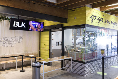 Photograph of a coffee shop. To the left of the shop is an outdoor seating area that is sectioned off with black stanchions. Above this seating area are two video monitors affixed to a wood beam. The left screen has the letters BLK in white against a black background, the right screen has a close crop on person's face looking upwards with their two arms raised upwards; there are purple lights behind them. The coffee shop exterior is split into three tiers, with a window into the cafe in the middle.