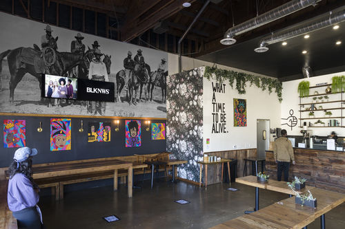 Image of the inside of coffee shop. One person in a sweater and baseball cap waits in line in the far left of the image and another person stands at the counter. To their left, a wall lined with five paintings. The wall behind the paintings is dark blue. Above that there is a wallpaper with an archival image of six black men on horses and one black man standing in front of them. Two video monitors are on top of the wallpaper. Left screen has four women standing together, right has words BLKNWS in white.